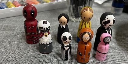 Målade träfigurer föreställande olika karaktärer står på ett bord med konstmaterial i bakgrunden.
