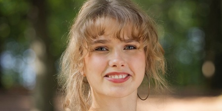 Person med ljust hår och vit blus står utomhus i en grönskande skogsmiljö.