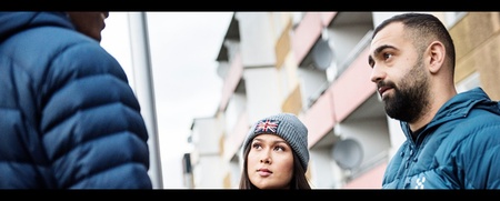 Tre personer samtalar utomhus framför ett bostadshus, två ansikten syns tydligt.