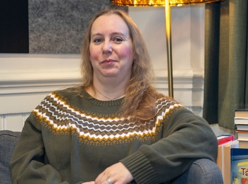 En kvinna med axellångt blont hår sitter i fåtölj med. Bakom henne syns en lampa och en trave böcker.