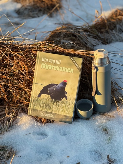 En bok med titeln 'Din väg till jägarexamen' och en termos med kopp står på snö vid torrt gräs i utomhusmiljö.