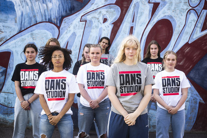En grupp unga personer står framför en graffitivägg, alla bär t-shirts med texten 'DANS STUDION'.