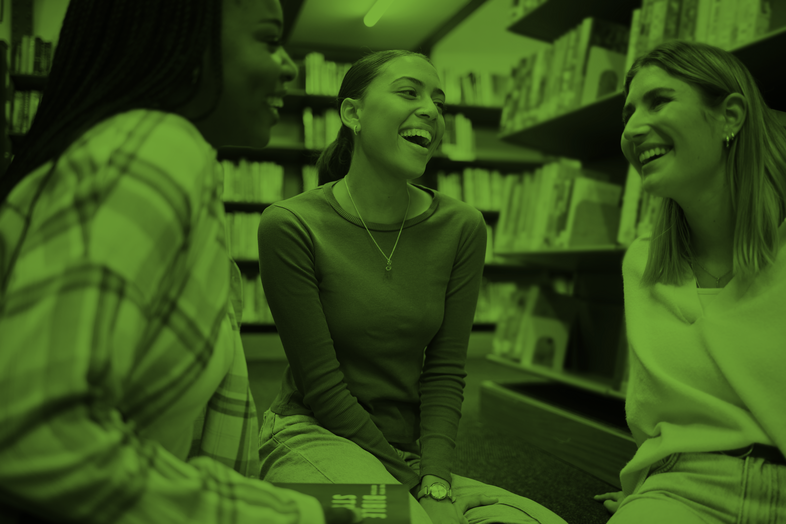 Tre unga kvinnor sitter och skrattar tillsammans i ett bibliotek.