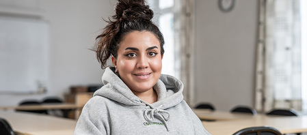 Ung kvinna med mörkt hår i knut och grå huvtröja sitter i ett klassrum och ler mot kameran.