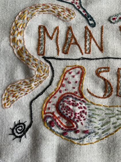 Broderi på tyg med texten 'MAN' och färgglada mönster och former.