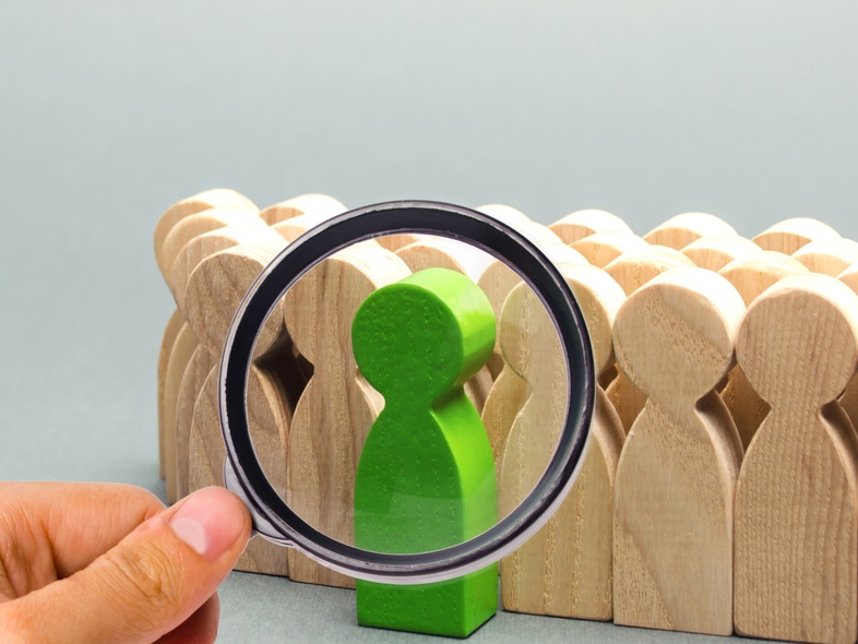En hand håller ett förstoringsglas över en grön träfigur bland flera beige träfigurer.