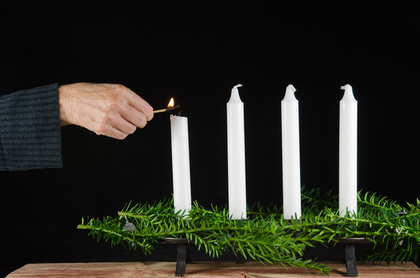En hand tänder det första av fyra vita ljus i en adventsljusstake med gröna kvistar.