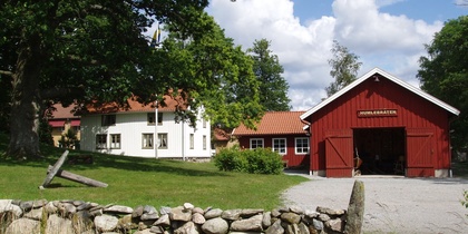 Gårdsmiljö med röd lada, vit byggnad, stengärdsgård och grönskande träd under blå himmel.