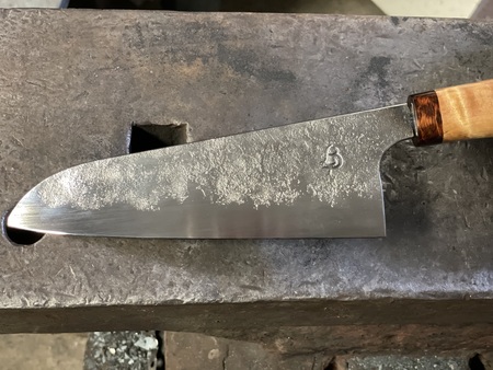 En handgjord kockkniv med trähandtag ligger på ett smidesstäd.