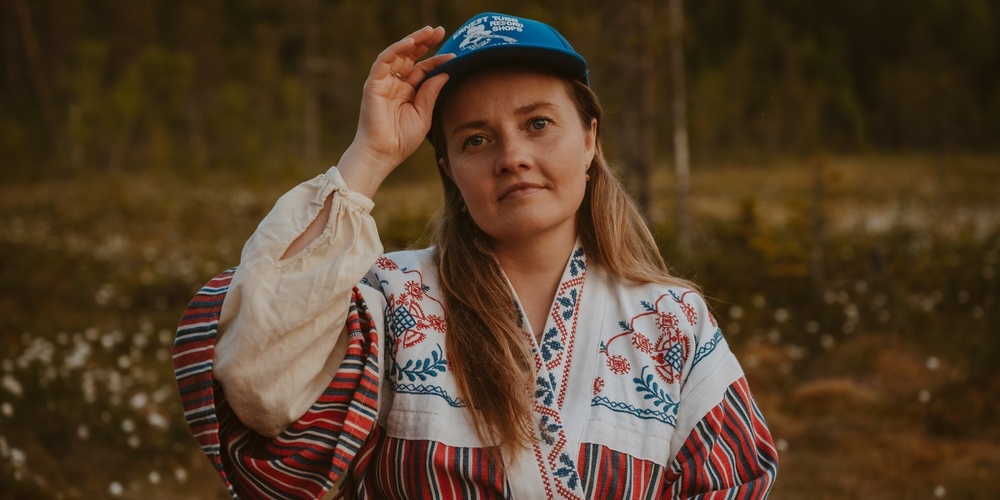 Person i traditionell broderad skjorta och blå keps står utomhus i naturmiljö.