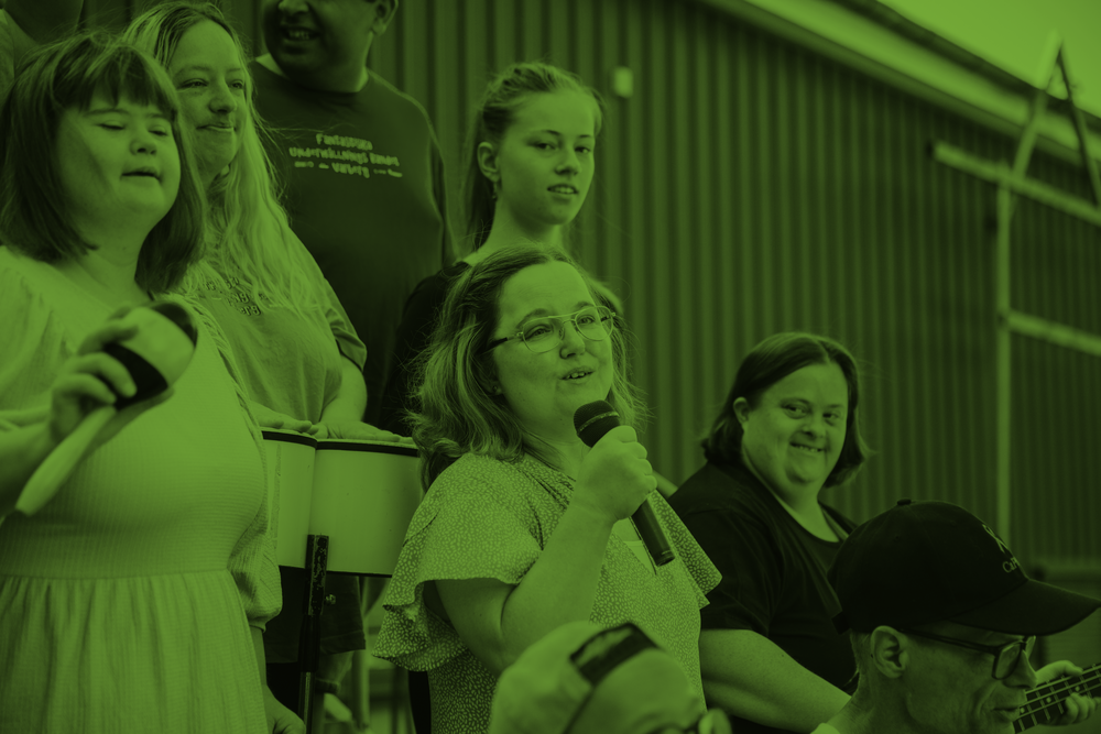 En grupp människor, varav en kvinna håller en mikrofon, sitter tillsammans inomhus. Bilden har grön färgton.