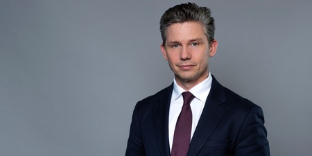 Man i mörk kostym och vinröd slips står mot en grå bakgrund.