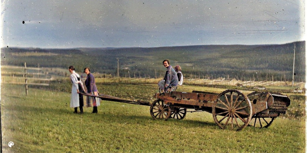 Två kvinnor drar en gammaldags trävagn med två personer sittande, på en äng med kullar i bakgrunden.