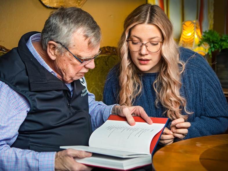 En äldre man och en yngre kvinna sitter tillsammans och läser i en bok i ett vardagsrum.