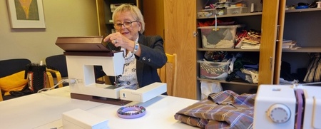 En äldre kvinna sitter vid ett bord och syr med en symaskin i ett rum med tyg och sybehör.