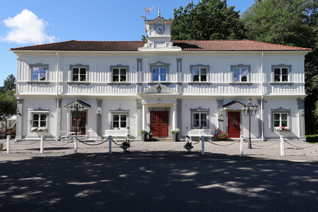 Vit tvåvånings herrgårdsbyggnad med rött tegeltak, symmetrisk fasad och klocktorn, omgiven av träd.