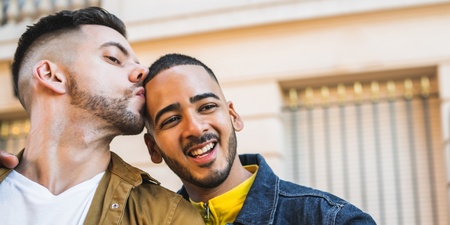 Två unga män tar en selfie utomhus, en kysser den andra på pannan och båda ser glada ut.