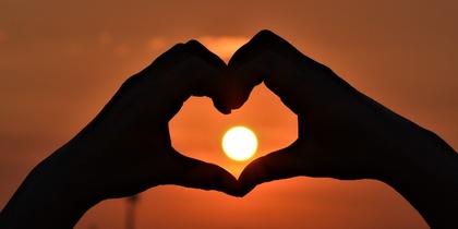 Två händer formar ett hjärta framför en solnedgång med solen i mitten av hjärtat.