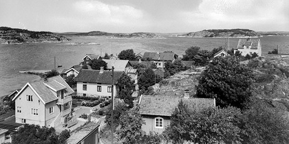 Svartvit bild av en liten by vid vattnet med flera hus, träd och berg i bakgrunden.
