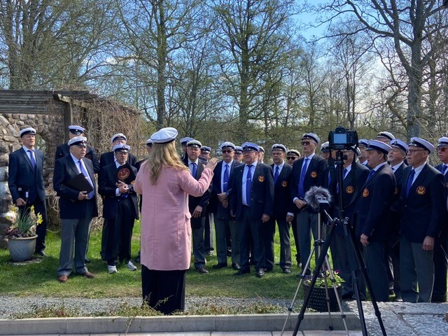 En grupp personer i kostym och studentmössor står utomhus, en kvinna dirigerar dem, kamera på stativ framför.