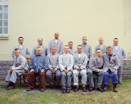 Gruppfoto av män i liknande kläder utomhus framför en ljus vägg, ansikten är suddade.