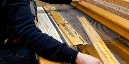 En person väver tyg på en stor vävstol i ett ljust rum. Händerna arbetar med trådarna.