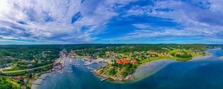 Panoramabild över en småstad vid vattnet med småbåtshamn, gröna skogar och blå himmel med moln.