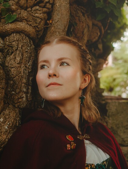 Person i medeltidsinspirerad klädsel står framför en gammal trädstam utomhus.