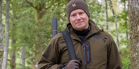 Man i brun jacka och mössa står i skog med gevär på axeln och ler mot kameran.