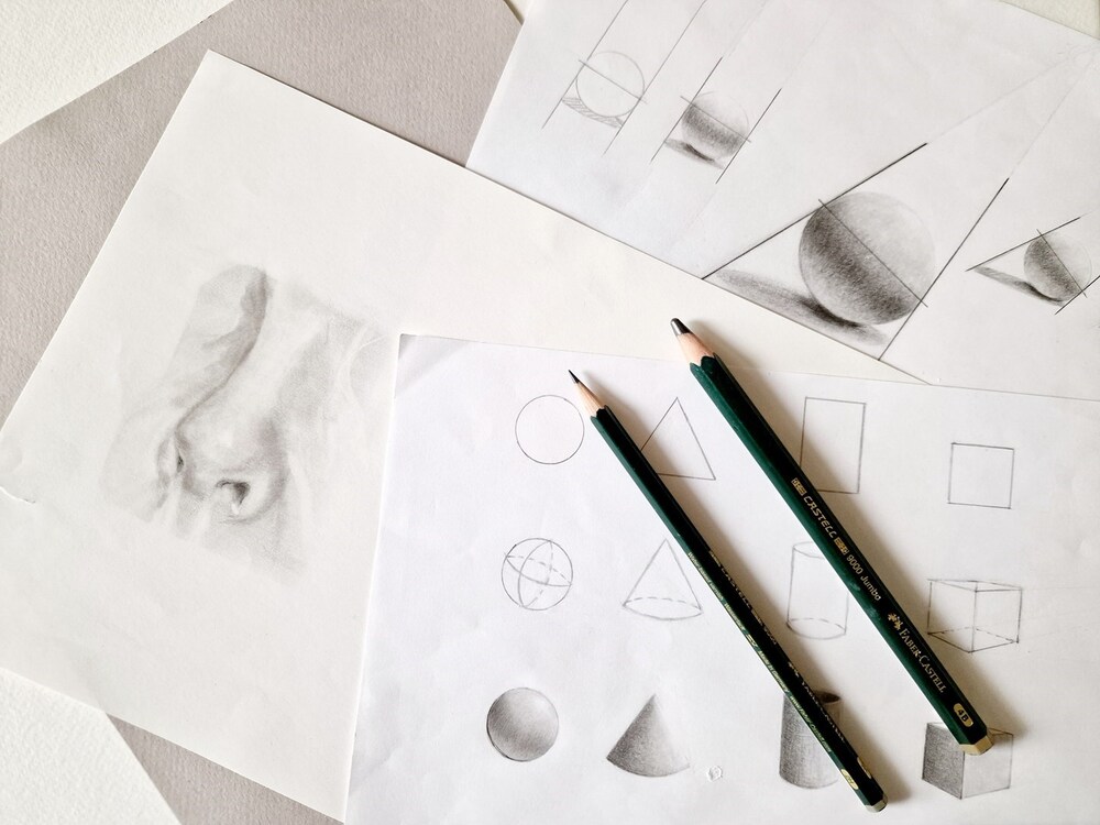 Flera papper med blyertsteckningar av geometriska former och en näsa, samt två pennor på ett bord.