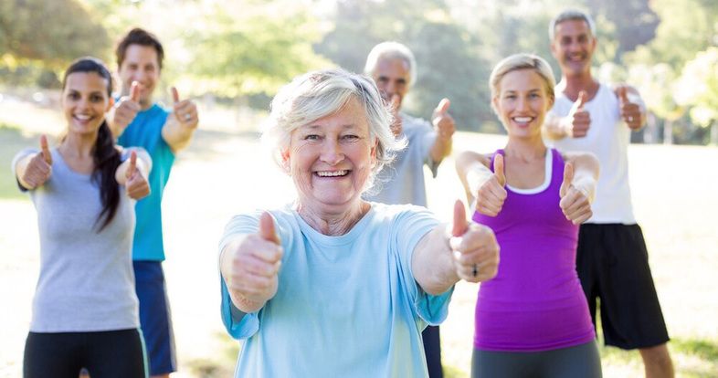 En grupp glada människor utomhus ger tummen upp mot kameran, med en äldre kvinna i fokus.