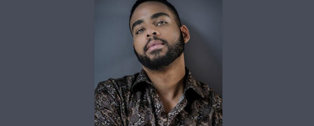 Man med kort skägg och mönstrad skjorta poserar mot en grå bakgrund.