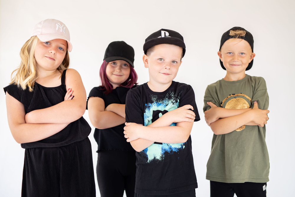 Fyra barn står i rad med armarna i kors och bär kepsar, mot en ljus bakgrund.
