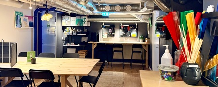 Ett modernt, färgglatt hobbyrum med arbetsbord, stolar, ventilationsrör i taket och pysselmaterial på bänkar.