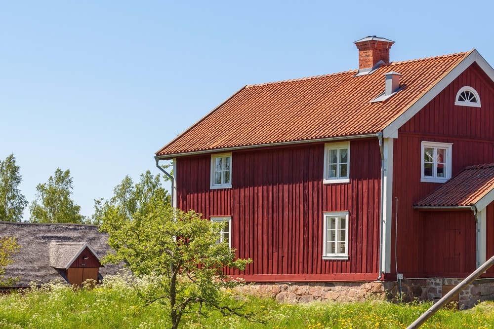 Två röda trähus med vita knutar på landet, omgivna av gräs, staket och blå himmel.