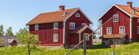 Två röda trähus med vita knutar på landet, omgivna av gräs, staket och blå himmel.