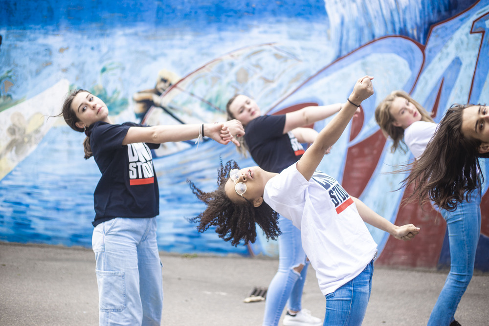 Fyra unga personer dansar energiskt framför en blå graffitivägg utomhus.