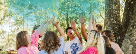 En grupp människor står i en ring utomhus och kastar färgat pulver i luften under träd.