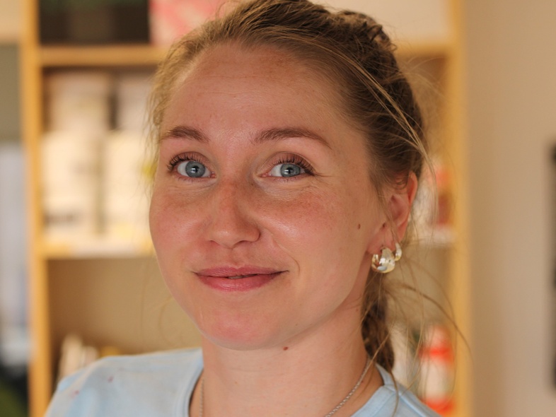 Kvinna med blont hår och blå ögon ler mot kameran i en inomhusmiljö.