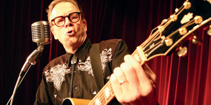 En person spelar gitarr och sjunger i mikrofon på scen med röda gardiner i bakgrunden.