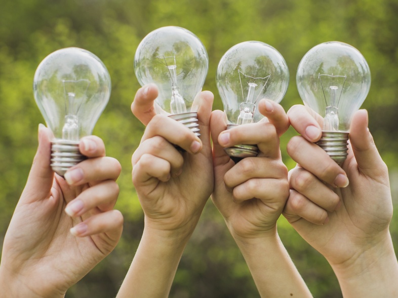 Fyra händer håller upp varsin glödlampa utomhus med grönskande bakgrund.