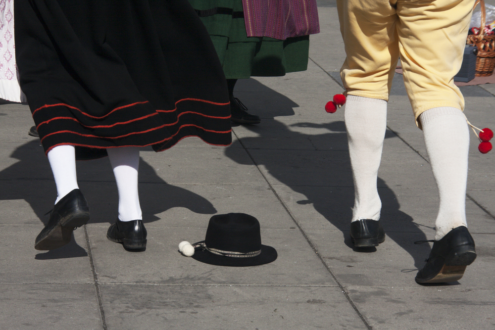 Två personer i traditionella folkdräkter dansar på en stenlagd yta, en svart hatt ligger på marken.