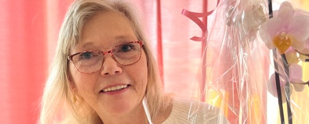 Leende kvinna med glasögon håller en inslagen orkidé framför röda gardiner.