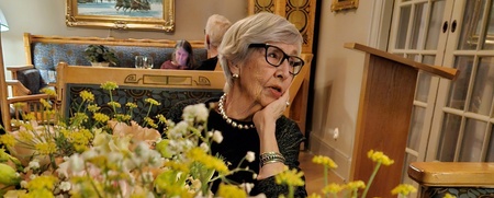 En kvinna med pärlhalsband sitter vid ett bord med blommor i ett elegant rum med konst på väggarna.