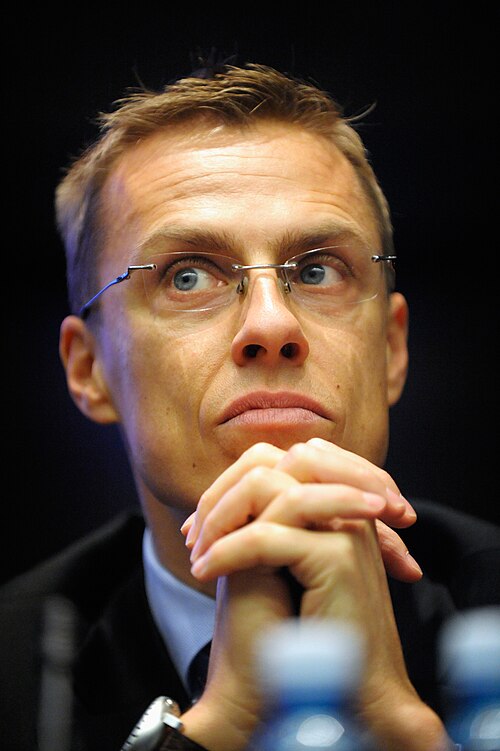 En person i kostym sitter med händerna knäppta framför sig vid ett bord med vattenflaskor.