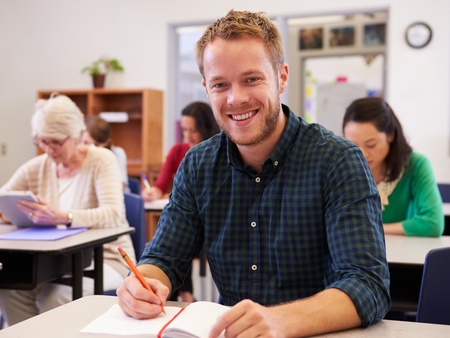 Leende man sitter vid skolbänk i klassrum, skriver i anteckningsbok. Andra vuxna studenter i bakgrunden.