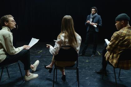 Fyra personer i ett mörkt rum, tre sitter med manus och en står framför dem, troligen under en repetition eller audition.