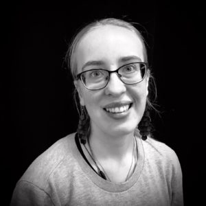 Person med ljust hår och grå tröja mot svart bakgrund.