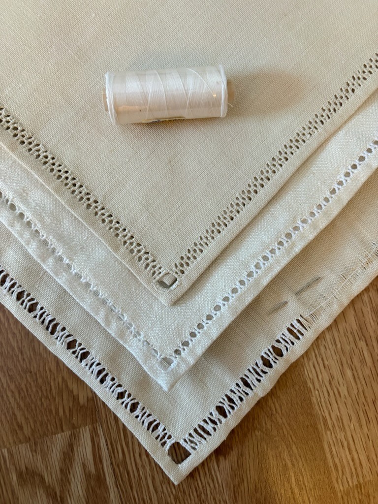 Tre beige linneservetter med dekorativa hålsömmar och en vit trådrulle ovanpå, liggande på ett trägolv.
