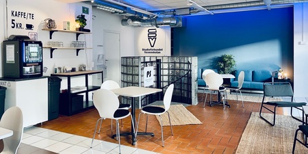Ett modernt café med vita stolar, små bord, kaffemaskin och blå vägg. Inga människor syns i lokalen.
